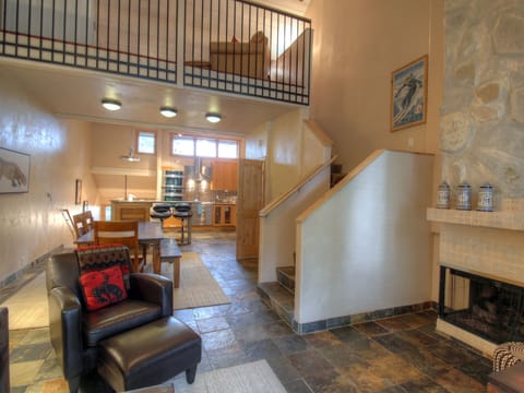 A cozy living area with a fireplace, black leather chair, hardwood flooring, an adjacent kitchen with wooden cabinetry, and an upstairs loft. A dining table and chairs are set near the kitchen.