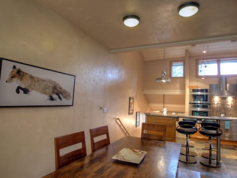A modern kitchen and dining area featuring wooden furniture, a painting of a fox on the wall, and contemporary kitchen appliances under natural and artificial lighting.
