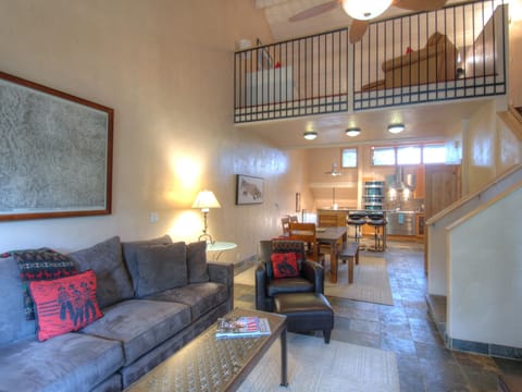 A modern living room with a gray couch, armchair, coffee table, and wall artwork. A loft area is seen above, and a dining table and kitchen are in the background. Stairs lead to the loft.