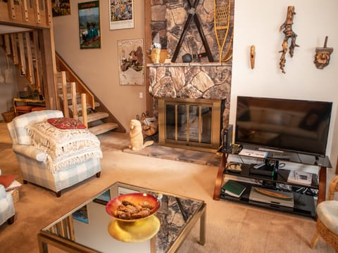 A cozy living room with a stone fireplace, TV, armchairs, and a glass coffee table. Stairs lead to an upper level, and the walls are adorned with various posters and masks.