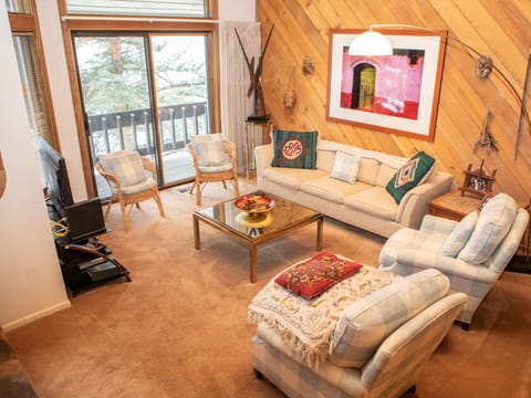 A cozy living room features beige sofas, a glass coffee table, and plaid armchairs. Art decorates a wood-paneled wall, while a sliding glass door leads to a snow-covered balcony outside.