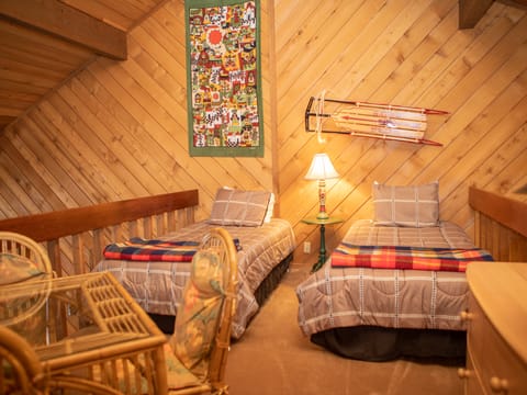 A cozy loft bedroom with two twin beds, a bedside table with a lamp, wooden walls, a wall hanging, vintage skis as decoration, and a wicker furniture set.