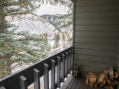 A small covered balcony with a stack of firewood overlooks snowy mountains and a pine tree. The balcony railing is painted dark gray.