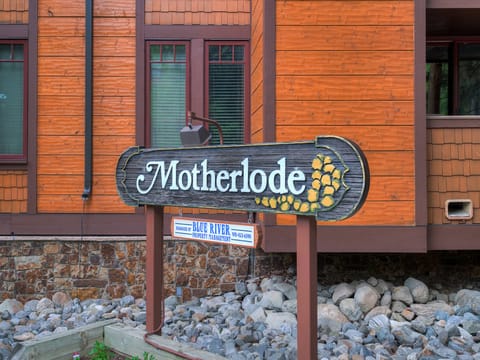 A wooden sign with the word "Motherlode" and a small "Blue River Property Management" sign below it. The signs are in front of a building with wooden siding and stone foundation.