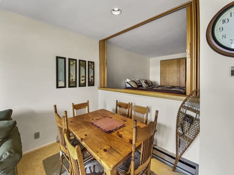 A dining area with a wooden table and chairs is shown. A window without glass reveals a bedroom with a bed. Framed art, a wall clock, and a snowshoe decor are visible on the walls.