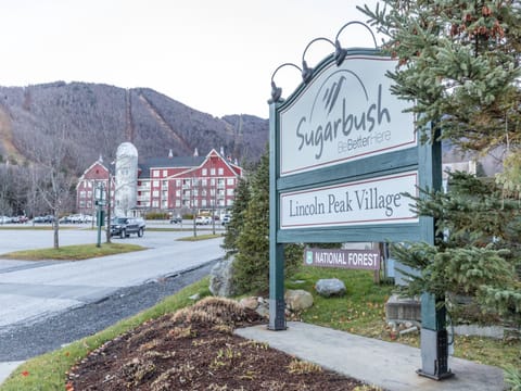 Entrance sign for Sugarbush Lincoln Peak Village, with visible parking lot, buildings, mountains in the background, and a National Forest sign at the base.