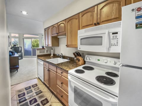 A narrow kitchen with wooden cabinets, a granite countertop, a white stove, a white microwave, and a white refrigerator. There is a sink, a dishwasher, and a rug on the tile floor. An open doorway leads to another room.