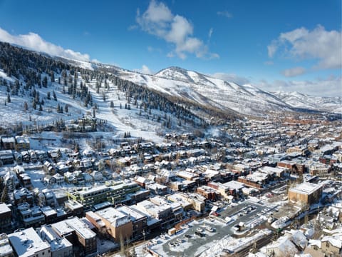 Aerial view of a snow-covered town with buildings, streets, and parked cars, set against a backdrop of tree-lined, snow-dusted mountains under a partly cloudy blue sky.