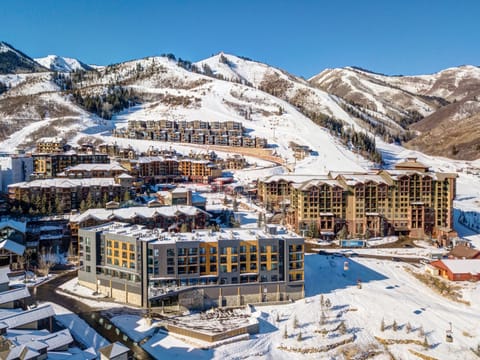 Aerial view of a snow-covered ski resort with multiple mid-rise buildings and surrounding mountain scenery on a clear day.