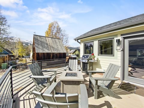 Outdoor patio with a fire pit surrounded by Adirondack chairs, a BBQ grill, and a vase of flowers on the corner of the railing. A barn and mountain landscape are visible in the background.