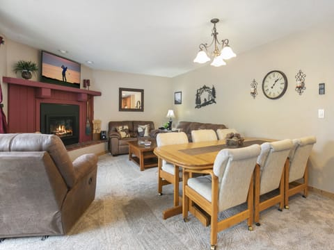A cozy living room features a fireplace, a flat-screen TV on the mantel, a brown sectional sofa, a recliner, a dining table with six chairs, and a wall clock. The carpeted floor adds to the warmth.