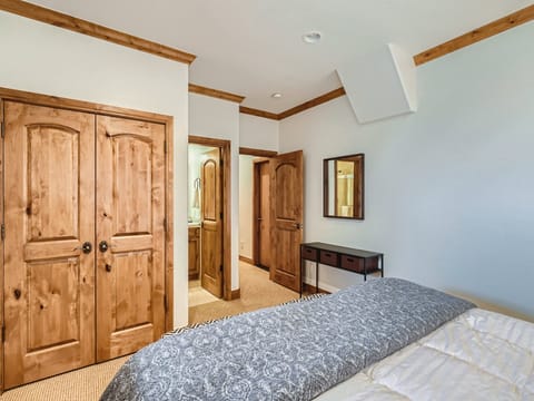 A bedroom with wood trim, a double-door closet, a bed with a patterned comforter, and a door leading to a bathroom. A small table with drawers is against the wall near the door.