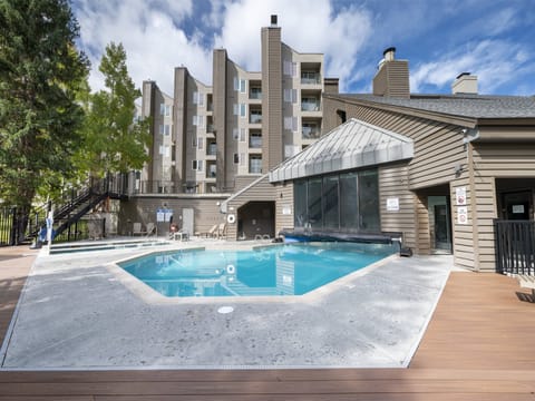 A modern condominium complex with multiple stories stands behind a fenced outdoor swimming pool, adjacent to a building with large windows, under a partly cloudy sky.
