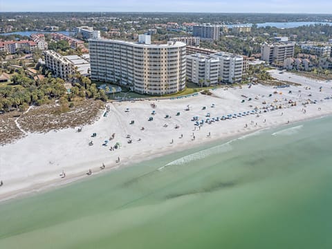 Aerial view of a coastal city featuring a wide sandy beach, ocean waves, beach chairs, umbrellas, and several mid-rise buildings and resorts.
