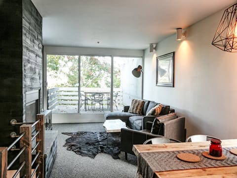 A modern living room with a gray sectional sofa, a mounted light fixture, a dining table, and a balcony view through large glass doors. There is a cowhide rug on the floor and a black accent wall.