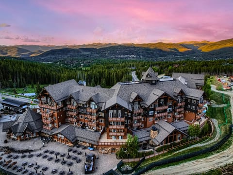 An aerial view of a large mountain lodge with multiple buildings, surrounded by dense forests and mountain peaks in the background at sunset.
