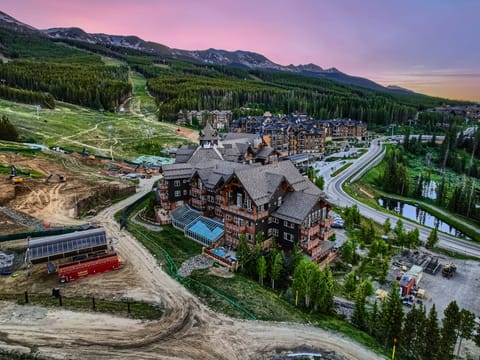Aerial view of a mountain resort complex with surrounding green hills and construction areas under a pink and blue sky at sunset. Roads and trees are visible in the scene.