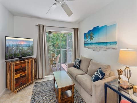 A cozy living room with a gray sofa, blue cushions, a wooden coffee table, a TV on a wooden stand, a ceiling fan, beach-themed artwork, and a sliding door leading to a balcony with outdoor seating.
