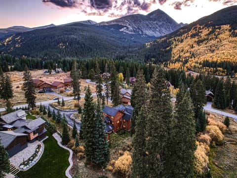 Aerial view of a mountainous landscape with houses and cabins nestled among tall trees and winding roads during autumn, with a backdrop of rugged peaks under a partly cloudy sky.