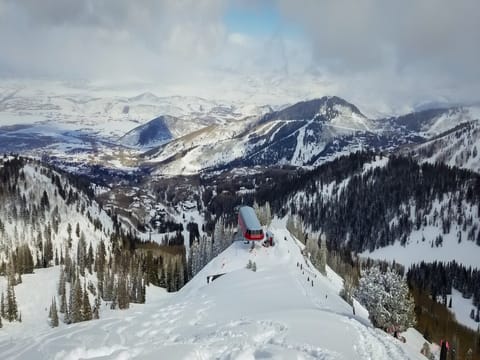 Snow-covered mountain landscape with a ski lift station at the center, surrounded by pine trees, with a sprawling valley and distant mountains under a partly cloudy sky.
