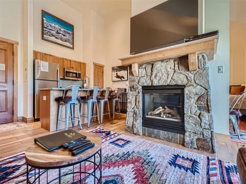 A cozy living space with a stone fireplace, flat-screen TV, and a wooden coffee table. The room features a modern kitchen with bar seating, colorful rugs, and mountain-themed wall art.