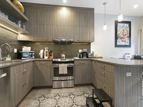 A modern L-shaped kitchen with wooden cabinets, stainless steel appliances, grey tiled backsplash, a patterned rug, and two pendant lights. A poster and small black stool are also visible.