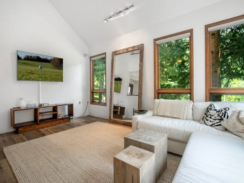 A modern living room with a white sectional sofa, a large wooden-framed mirror, a TV mounted on the wall, and large windows offering a view of greenery. The decor is minimalist with wooden accents.
