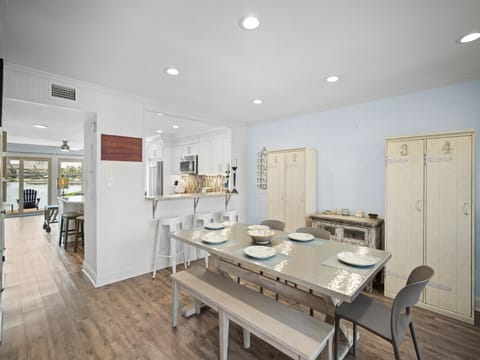 Bright dining area with seating for six, barstools at the kitchen counter, and LVP wood floors. Features a clean coastal style with light furnishings and an open layout.