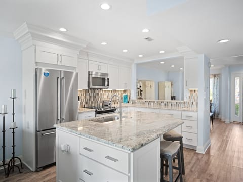 Modern kitchen with a large granite island, stainless steel appliances, white shaker cabinets, tile backsplash, and recessed lighting. Features include bar seating, hardwood floors, and a mirrored wall for added depth.