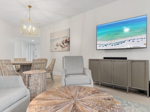 This light-filled coastal living area features soft neutral tones, textured wood furnishings, and beachy artwork. A smart TV and comfy seating invite relaxation, while the open layout flows effortlessly into the dining space for easy entertaining.