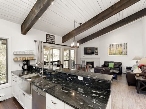 A modern kitchen with a black granite countertop, stainless steel appliances, and an open view into a living area with wooden beams on the ceiling and a fireplace.