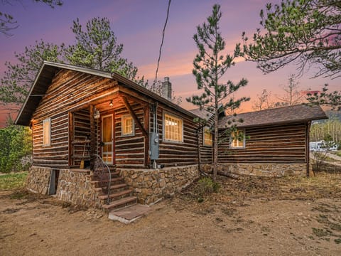 Rustic log cabin with stone foundation nestled among evergreen trees, photographed during twilight hours with dramatic purple-orange sky.