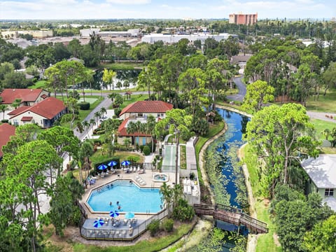 Aerial view of a vacation resort featuring an outdoor pool, tree-lined canal, and residential buildings set within a lush tropical landscape.