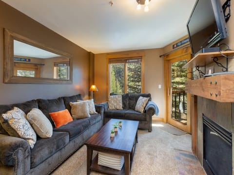 A living room with two gray couches, a wooden coffee table, a TV mounted above a fireplace, and large windows with a forest view. Pillows are arranged on the couches, and a plant decorates the table.