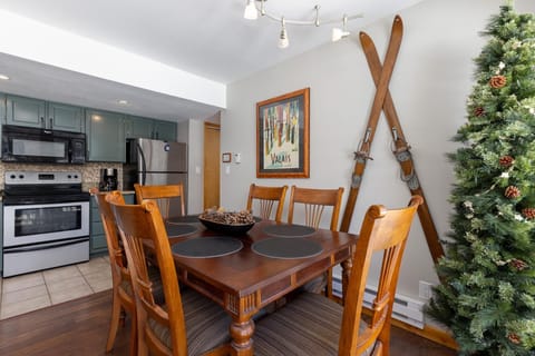 A dining area with a wooden table set for four, a kitchen in the background, and a decorated Christmas tree. Antique wooden skis are mounted on the wall, and a framed poster hangs above them.