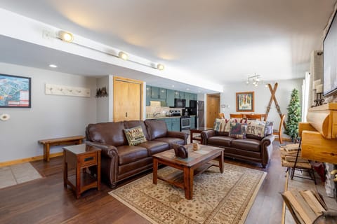 A cozy living room with two brown leather sofas, a wooden coffee table, and a patterned rug. The background features a kitchen with a breakfast bar and wooden cabinets. Walls are adorned with decor.