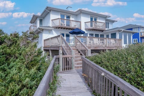 Charming beachside home with wooden walkway leading to elevated decks and multiple balconies overlooking coastal vegetation.