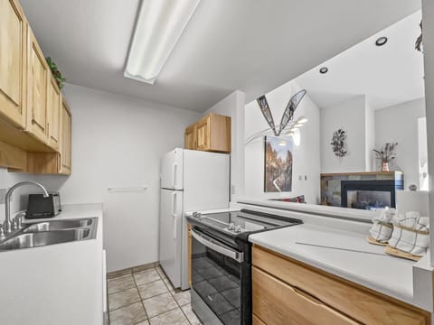 A small kitchen featuring wooden cabinets, a white refrigerator, and an electric stove. The kitchen opens to a living area with a wall-mounted TV and a ceiling fan with leaf-shaped blades.