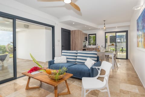 A bright contemporary living room featuring a blue sofa with white stripes, a wooden coffee table adorned with plants and fruits, and an open kitchen in the background. Artistic touches like a colorful painting add to the warm ambiance.