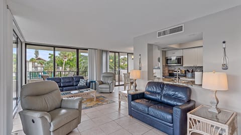 Modern living room with tiled floor, featuring two leather sofas, two recliner chairs, and a glass coffee table. The room has large windows, a view of a balcony, and an adjacent open kitchen area.