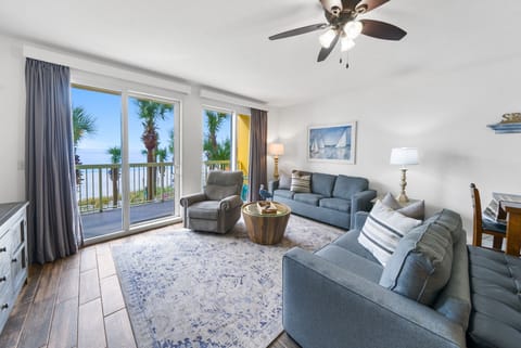 Relax in your ocean-view living room with comfortable seating, gentle ceiling fan, and sliding doors opening to palm-lined balcony.