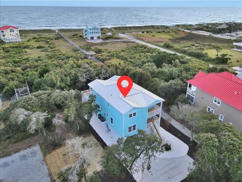 Aerial view of a bright blue beach house with a red location pin, porches, driveway, trees, dunes, a beach boardwalk, nearby homes, and Atlantic Ocean views in Oak Island, NC.