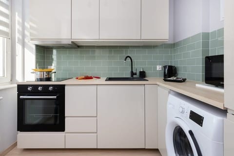 A compact kitchen with white cabinetry, a light-green tiled backsplash, and essential appliances including an oven and washing machine.

