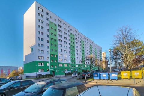 An exterior shot of a tall residential building painted in pastel colors, with parked cars and a tree-lined sidewalk in front.







