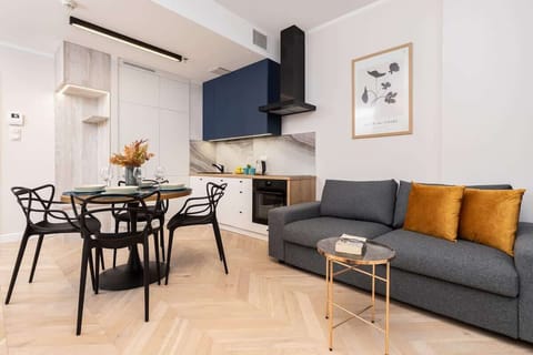 A modern living and dining area featuring a grey sofa with mustard-yellow cushions, a round dining table with black chairs, and large windows with gray curtains.

