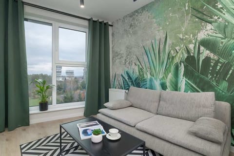 A bright living area featuring a large grey sofa set against a striking botanical wallpaper, with floor-to-ceiling windows and green curtains.

