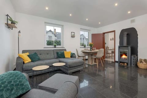A spacious living room with glossy dark flooring, a large grey corner sofa accented by green and yellow pillows, and natural light streaming in through multiple windows.