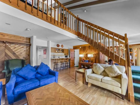 A spacious living area featuring wooden accents, a staircase with railings leading to an upper level, two sofas, a coffee table, and a breakfast bar with stools. The room has a cozy, rustic feel.