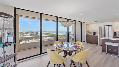 A modern dining area with a glass table set with yellow chairs, overlooking a coastal view through large floor-to-ceiling windows. The adjacent kitchen features stainless steel appliances and a central island.