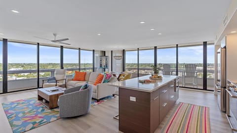 Modern open-plan living room and kitchen with floor-to-ceiling windows, contemporary furniture, colorful rugs, and a cityscape view. The room is filled with natural light.